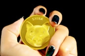 A hand with dark nail polish holds a golden coin featuring an embossed image of a Shiba Inu dog and the word 'SHIBA' at the top. The focus is primarily on the coin, with a black background enhancing its prominence.