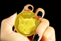 A hand with dark nail polish holds a golden coin featuring an embossed image of a Shiba Inu dog and the word 'SHIBA' at the top. The focus is primarily on the coin, with a black background enhancing its prominence.