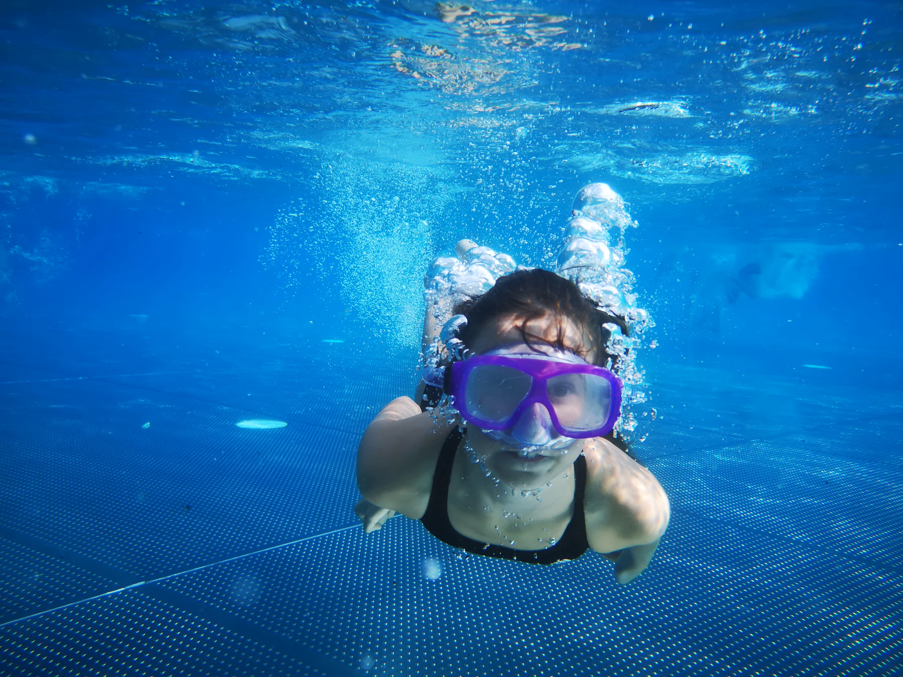 A child in goggles swimming underwater in a bright blue pool