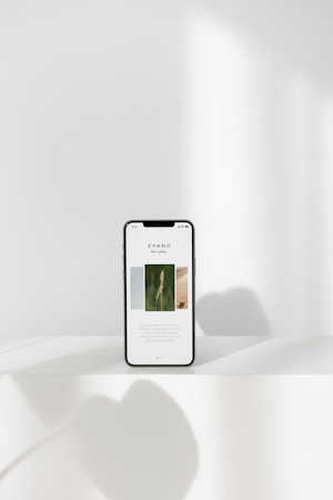 A smartphone displaying a minimalist webpage stands on a white surface, with soft shadows cast around it. The background is a pale, neutral tone with subtle lighting.