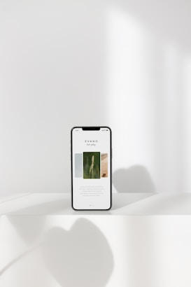 A smartphone displaying a minimalist webpage stands on a white surface, with soft shadows cast around it. The background is a pale, neutral tone with subtle lighting.