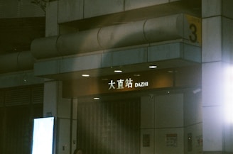 An entrance to a metro station labeled 'DAZHI' with Chinese characters. The setting appears dimly lit, possibly during nighttime, with a spotlight illuminating the station sign. There is a number '3' on a yellow sign, indicating the entrance or exit number.