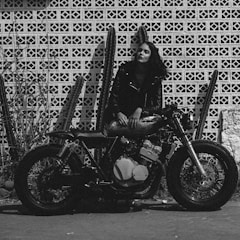 A woman in a leather jacket is leaning on a vintage motorcycle. The background features a patterned brick wall with several tall cacti. The scene is rendered in black and white, giving it a gritty, classic look.
