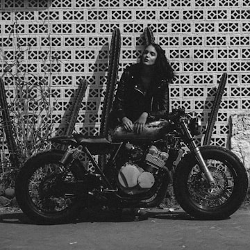 A woman in a leather jacket is leaning on a vintage motorcycle. The background features a patterned brick wall with several tall cacti. The scene is rendered in black and white, giving it a gritty, classic look.