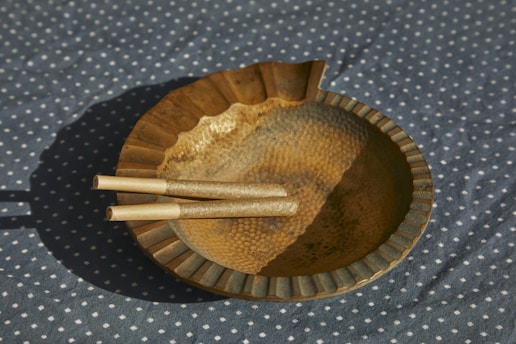 A round, metallic ashtray with a textured, hammered surface and scalloped edges is placed on a blue polka dot fabric. The ashtray contains three unlit, rolled cigarettes.