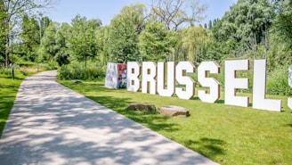 Large white letters spell out 'BRUSSELS' on a grassy area next to a walking path. The path is surrounded by lush green trees and shrubs under a bright blue sky. Stones are scattered along the grass, adding to the park-like setting.
