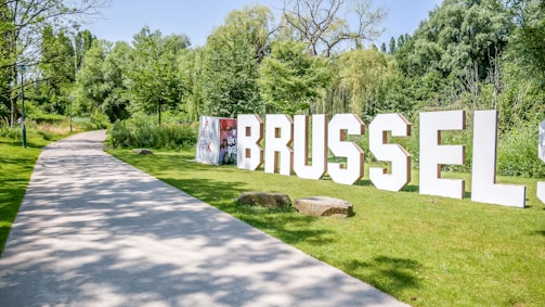 Large white letters spell out 'BRUSSELS' on a grassy area next to a walking path. The path is surrounded by lush green trees and shrubs under a bright blue sky. Stones are scattered along the grass, adding to the park-like setting.