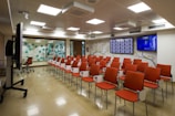 A modern conference room with rows of orange padded chairs arranged in facing a wall with two large flat screens, one showing multiple profiles or icons and the other broadcasting a show or presentation. The room has a polished floor, fluorescent ceiling lights, and a decor that includes a floral branch mural and a wall of hexagonal patterns in teal and white. A large monitor on a stand is positioned to the side.