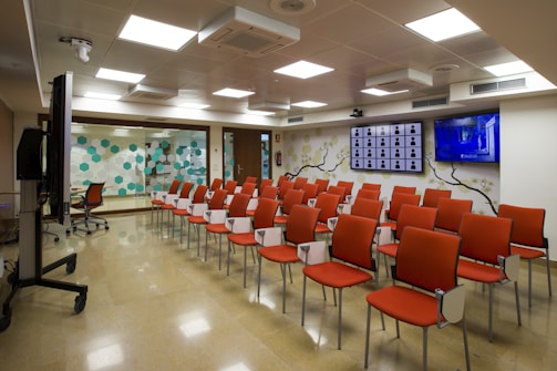 A modern conference room with rows of orange padded chairs arranged in facing a wall with two large flat screens, one showing multiple profiles or icons and the other broadcasting a show or presentation. The room has a polished floor, fluorescent ceiling lights, and a decor that includes a floral branch mural and a wall of hexagonal patterns in teal and white. A large monitor on a stand is positioned to the side.