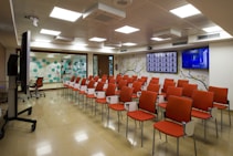 A modern conference room with rows of orange padded chairs arranged in facing a wall with two large flat screens, one showing multiple profiles or icons and the other broadcasting a show or presentation. The room has a polished floor, fluorescent ceiling lights, and a decor that includes a floral branch mural and a wall of hexagonal patterns in teal and white. A large monitor on a stand is positioned to the side.