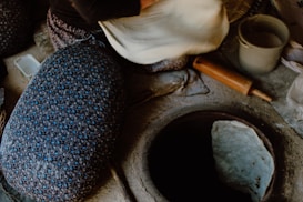 A person is preparing dough near a traditional tandoor oven. The scene includes a rolling pin, a patterned cloth, a container, and a raw piece of dough on the counter.