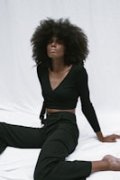 A person with voluminous curly hair wearing a black outfit is seated on a white backdrop, striking a relaxed pose.