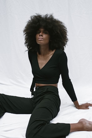 A person with voluminous curly hair wearing a black outfit is seated on a white backdrop, striking a relaxed pose.