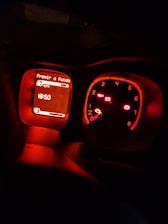 A car dashboard showing illuminated dials and indicators in a dimly lit setting. The left display screen has text in Portuguese, likely indicating a mode or instruction, and shows a digital clock displaying 18:50. The speedometer and RPM gauge are lit up in red hues.