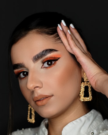 A close-up portrait of a person with striking makeup and large gold earrings. The individual has bold orange and black eye makeup, perfectly manicured nails, and is wearing a white outfit with pearl details. The hairstyle is sleek and pulled back with a focus on the facial features.