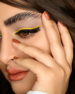 A close-up shot of a person's face showing detailed makeup with a bold yellow eyeliner, arched eyebrows, and terracotta-colored lips. The image focuses on the eye, highlighting the eyelashes and the precise makeup application. The person's fingers rest gently on the face, showcasing neatly manicured nails that match the lip color.