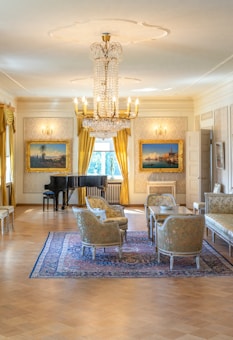 A luxurious and elegantly furnished room featuring a grand chandelier, vintage paintings in ornate frames on the walls, a black grand piano, golden curtains, and a set of richly upholstered chairs and sofas around a wooden table on an intricate area rug.