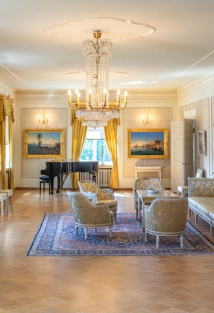 A luxurious and elegantly furnished room featuring a grand chandelier, vintage paintings in ornate frames on the walls, a black grand piano, golden curtains, and a set of richly upholstered chairs and sofas around a wooden table on an intricate area rug.