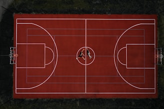 An aerial view of a red basketball court with two people lying at the center. The court is well-marked with lines for basketball play, and it is surrounded by a fence. The surrounding area is dark, creating a strong contrast with the vivid red of the court.
