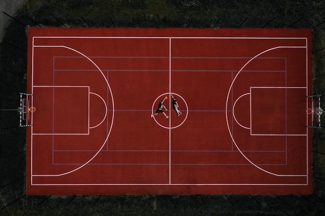 An aerial view of a red basketball court with two people lying at the center. The court is well-marked with lines for basketball play, and it is surrounded by a fence. The surrounding area is dark, creating a strong contrast with the vivid red of the court.