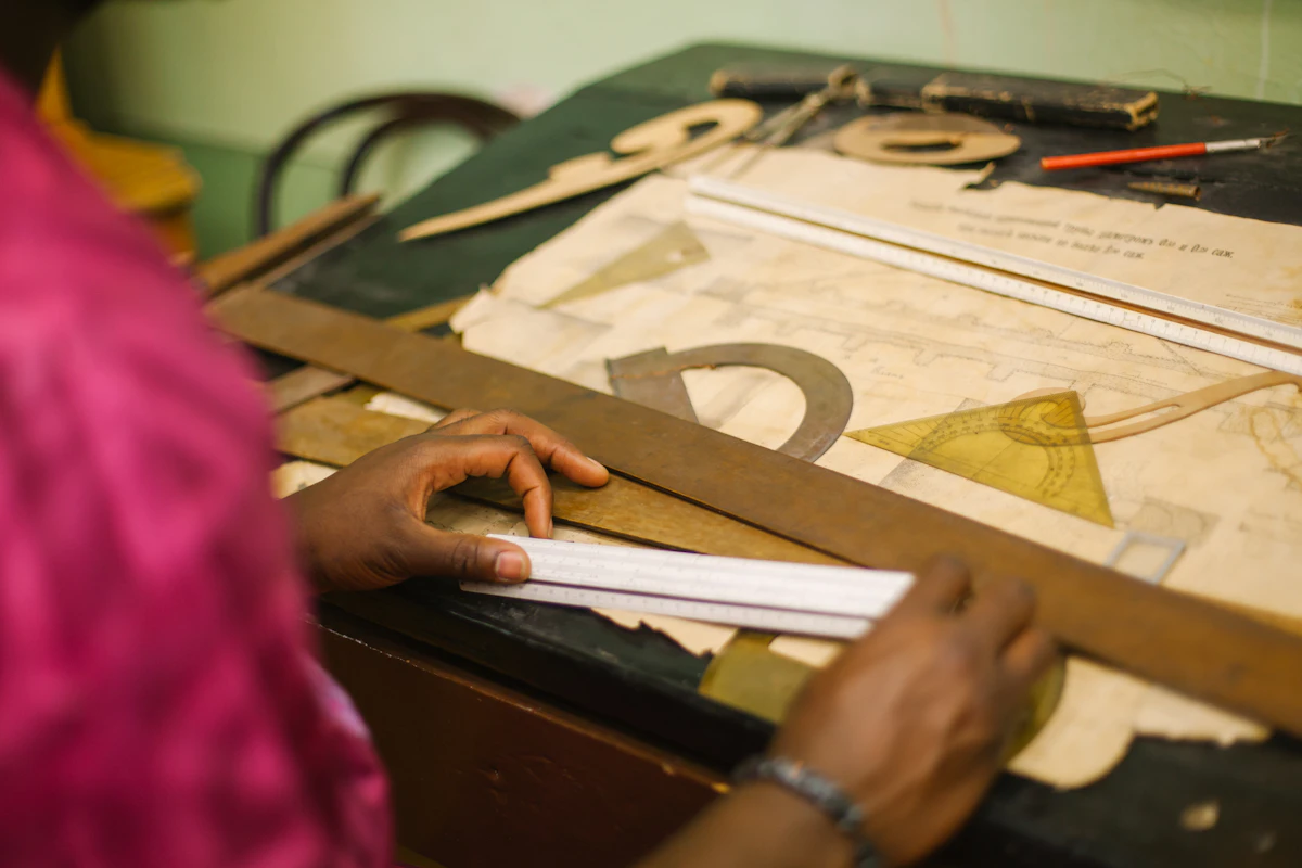 Two hands steadying a steel rule on a drafting board beside a set square and a french curve