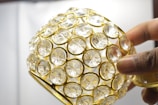 A hand holding a decorative object covered with clear, circular crystals set in a golden lattice framework.