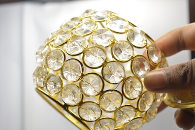 A hand holding a decorative object covered with clear, circular crystals set in a golden lattice framework.