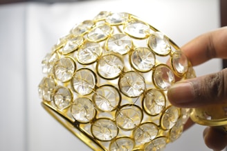 A hand holding a decorative object covered with clear, circular crystals set in a golden lattice framework.