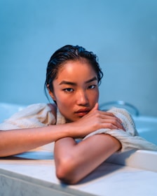 A person with wet hair reclines by the edge of a bathtub, resting their chin on crossed arms. The setting has a calming atmosphere with a blue-toned background and the subject's expression is contemplative.