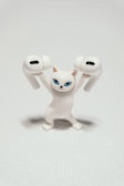 A small, white figurine of a cat with blue eyes is holding two wireless earbuds. The cat's expression is serious and determined, creating a humorous juxtaposition with its small size and the large earbuds.