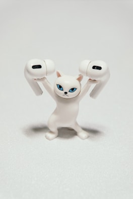 A small, white figurine of a cat with blue eyes is holding two wireless earbuds. The cat's expression is serious and determined, creating a humorous juxtaposition with its small size and the large earbuds.