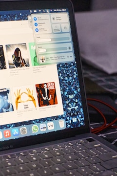 A laptop displaying a music player interface, showing album covers and song details. A control panel is open, allowing access to settings like Wi-Fi and Bluetooth. The screen has a stone-patterned wallpaper, and various app icons are visible on the dock. The laptop is an Apple product, as indicated by the design of the keyboard and interface.