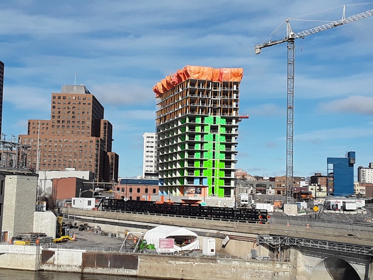 A construction site featuring a high-rise building under development with distinct green and orange sections. A tall crane towers to the right of the structure. Surrounding the construction are several other buildings of varying heights and designs, with a clear blue sky in the background.