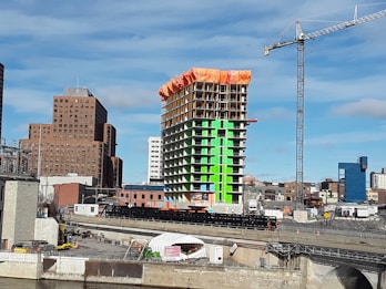A construction site featuring a high-rise building under development with distinct green and orange sections. A tall crane towers to the right of the structure. Surrounding the construction are several other buildings of varying heights and designs, with a clear blue sky in the background.