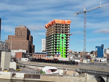 A construction site featuring a high-rise building under development with distinct green and orange sections. A tall crane towers to the right of the structure. Surrounding the construction are several other buildings of varying heights and designs, with a clear blue sky in the background.