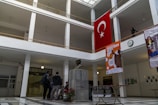 A spacious indoor hallway with a high ceiling and large windows allowing natural light to illuminate the area. A bust of a person is centrally placed on a marble pedestal in the foreground. The walls feature several posters and a large Turkish flag is prominently displayed on the upper floor. The floor is tiled in a geometric pattern, and several people are gathered near the bust, engaged in conversation.