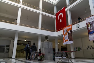 A spacious indoor hallway with a high ceiling and large windows allowing natural light to illuminate the area. A bust of a person is centrally placed on a marble pedestal in the foreground. The walls feature several posters and a large Turkish flag is prominently displayed on the upper floor. The floor is tiled in a geometric pattern, and several people are gathered near the bust, engaged in conversation.