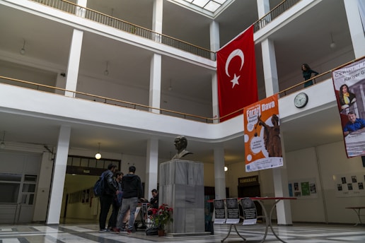A spacious indoor hallway with a high ceiling and large windows allowing natural light to illuminate the area. A bust of a person is centrally placed on a marble pedestal in the foreground. The walls feature several posters and a large Turkish flag is prominently displayed on the upper floor. The floor is tiled in a geometric pattern, and several people are gathered near the bust, engaged in conversation.