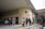 A group of people stand outside a university building with the sign 'Ankara Üniversitesi DİL ve TARİH - COĞRAFYA FAKÜLTESİ' on a stone facade. The entrance features a series of windows and a flat canopy. Some individuals are engaged in conversation while others appear to be using mobile devices. Trees without leaves and overcast sky indicate a colder season.
