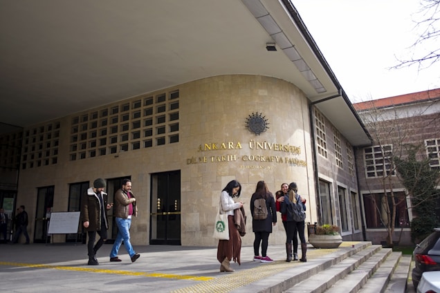 A group of people stand outside a university building with the sign 'Ankara Üniversitesi DİL ve TARİH - COĞRAFYA FAKÜLTESİ' on a stone facade. The entrance features a series of windows and a flat canopy. Some individuals are engaged in conversation while others appear to be using mobile devices. Trees without leaves and overcast sky indicate a colder season.