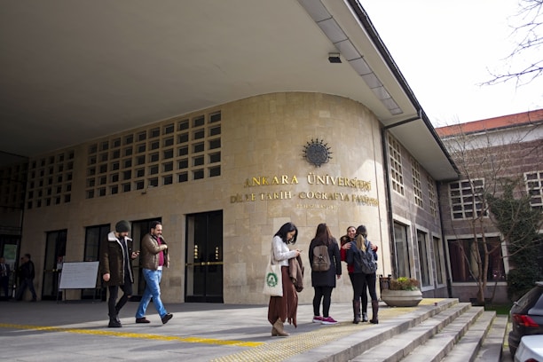 A group of people stand outside a university building with the sign 'Ankara Üniversitesi DİL ve TARİH - COĞRAFYA FAKÜLTESİ' on a stone facade. The entrance features a series of windows and a flat canopy. Some individuals are engaged in conversation while others appear to be using mobile devices. Trees without leaves and overcast sky indicate a colder season.
