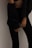 A person wearing dark clothing is standing against a plain wall. The outfit consists of fitted pants and a loosely buttoned shirt, partially revealing an undergarment. The focus is on the lower torso and legs.