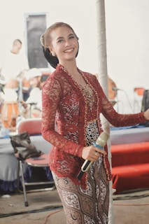 A woman dressed in an intricate maroon and gold traditional outfit holds a microphone. She has a joyful expression with her hair neatly styled. In the background, blurred figures and musical instruments suggest a performance setting.
