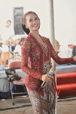 A woman dressed in an intricate maroon and gold traditional outfit holds a microphone. She has a joyful expression with her hair neatly styled. In the background, blurred figures and musical instruments suggest a performance setting.