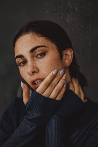 A person with wet hair and droplets on their face, wearing a dark sweater and showcasing colorful painted nails. The expression is thoughtful, and the pose suggests introspection, with hands gently touching the face.