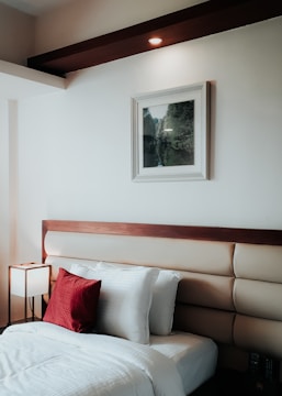 A neatly arranged bedroom features a stylish headboard with a wooden accent and padded upholstery. The white bedsheets create a crisp, clean look, accented by a vibrant red throw pillow. A small, modern lamp with a square shade is placed on a bedside table, providing soft lighting. A framed picture with a landscape scene hangs on the wall above the headboard. The overall decor is minimalistic and warm.
