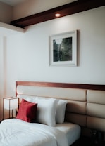 A neatly arranged bedroom features a stylish headboard with a wooden accent and padded upholstery. The white bedsheets create a crisp, clean look, accented by a vibrant red throw pillow. A small, modern lamp with a square shade is placed on a bedside table, providing soft lighting. A framed picture with a landscape scene hangs on the wall above the headboard. The overall decor is minimalistic and warm.