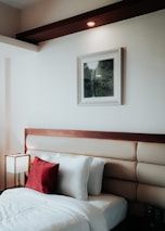 A neatly arranged bedroom features a stylish headboard with a wooden accent and padded upholstery. The white bedsheets create a crisp, clean look, accented by a vibrant red throw pillow. A small, modern lamp with a square shade is placed on a bedside table, providing soft lighting. A framed picture with a landscape scene hangs on the wall above the headboard. The overall decor is minimalistic and warm.