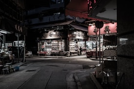 A dimly lit urban street scene featuring a store with illuminated displays filled with various goods. The street appears quiet with minimal people, and there are numerous signs with Chinese characters. The mood is moody, with a contrast between the dark surroundings and the bright storefront.