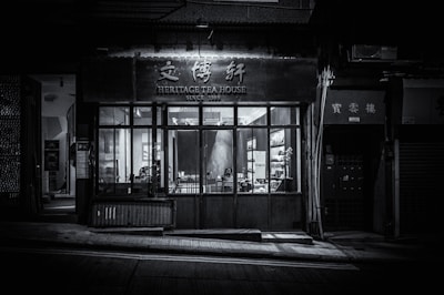 A dimly lit, monochrome exterior of a tea house with a sign that reads 'Heritage Tea House' above the entrance. The large front windows display part of the interior, showing tables and chairs. The establishment appears closed, with minimal light and no visible patrons.